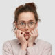 unhappy-nervous-young-female-with-worried-expression-bites-finger-nails-looks-anxiously-directly-wears-spectacles-dressed-fashionable-clothes-stands-against-white-wall