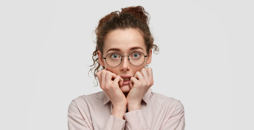 unhappy-nervous-young-female-with-worried-expression-bites-finger-nails-looks-anxiously-directly-wears-spectacles-dressed-fashionable-clothes-stands-against-white-wall
