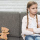 portrait-sad-girl-with-crossed-arms-sitting-near-teddy-bear-gray-sofa