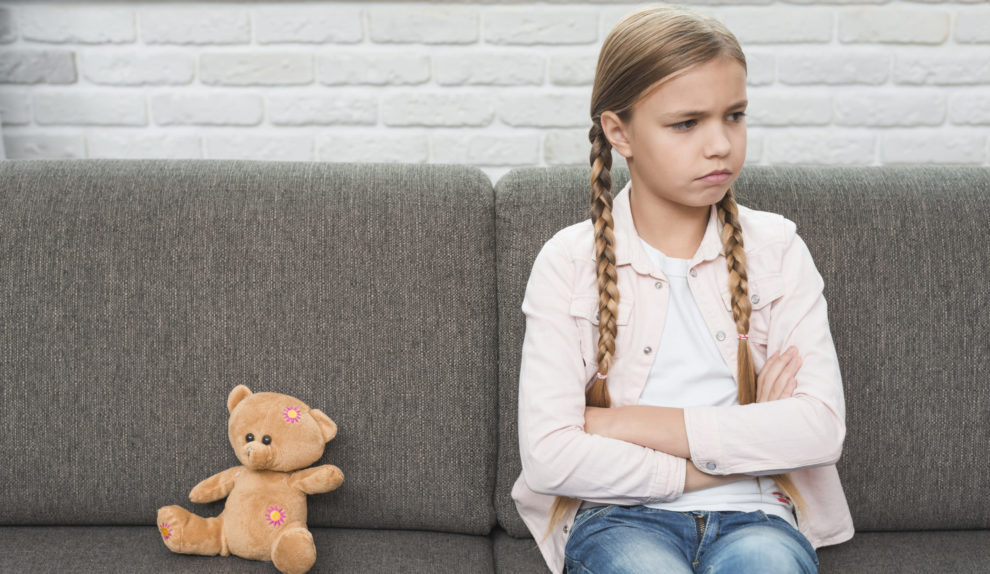 portrait-sad-girl-with-crossed-arms-sitting-near-teddy-bear-gray-sofa