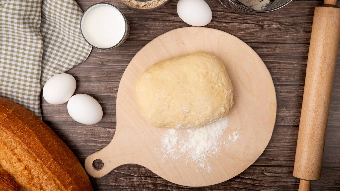 top-view-dough-with-flour-cutting-board-eggs-milk-baguette-rolling-pin-wooden-background_141793-5788